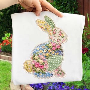 以下が含まれることがあります： カラフルなパッチワークのウサギのデザインが施された白い生地。ウサギは、ピンクのバラの刺繍と真珠のビーズの輪郭を持つ、花柄の生地のピースで作られています。デザインは手で持たれています。