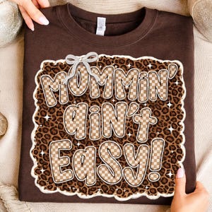 Puede incluir: Una sudadera marrón con la frase "Mommin' Ain't Easy!" en una fuente decorativa. El texto está delineado en blanco y relleno con un patrón de cuadros. El fondo es un estampado de leopardo.