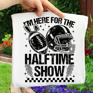 May include: White tote bag with a black graphic featuring a football with bunny ears, a football helmet, and the text "I'M HERE FOR THE HALFTIME SHOW". The design includes stars and checkered lightning bolts.