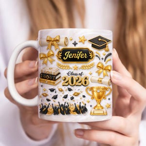 Puede incluir: Taza de cerámica blanca con temática de graduación. La taza presenta detalles dorados, incluyendo lazos, estrellas, un birrete y un trofeo. También se muestra el nombre "Jenifer" y el texto "Class of 2026".