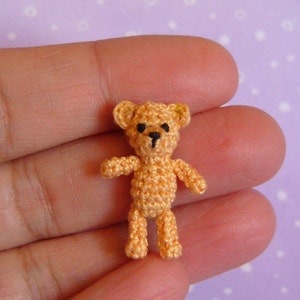 May include: A small crocheted teddy bear with brown eyes and a black nose. The bear is light orange in color and is standing on a person's hand.