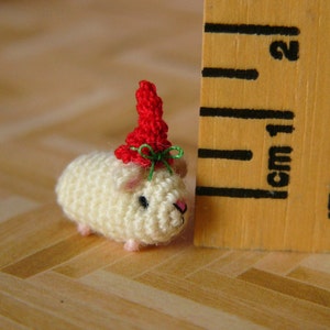 May include: A small crocheted hamster wearing a red and green hat. The hamster is about 1 inch tall and is sitting on a wooden surface.