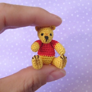 May include: A small crocheted Winnie the Pooh bear wearing a red shirt. The bear is sitting on a finger.