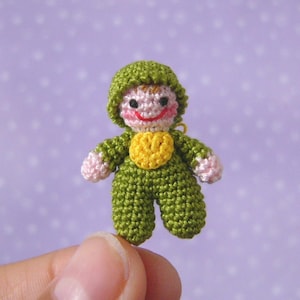 May include: A small crocheted doll wearing a green outfit with a yellow heart on the chest. The doll has a pink face with black eyes and a red smile.