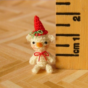 May include: A small crocheted teddy bear wearing a red and green hat. The bear is about 1 inch tall and is standing next to a ruler.