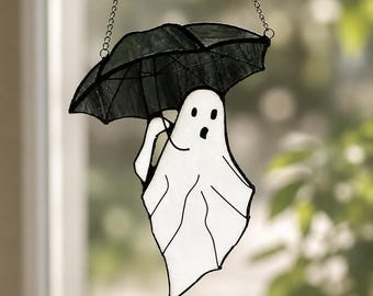May include: A stained glass suncatcher featuring a white ghost figure holding a black umbrella. The ghost has two black eyes and an open mouth. The piece is constructed with dark metal solder lines and includes a silver chain for hanging.