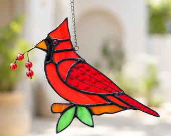 May include: A stained glass suncatcher shaped like a red cardinal bird perched on an orange branch with green leaves. The bird holds a small twig with three red glass beads in its beak. It is suspended by a thin metal chain for hanging display.