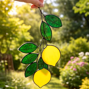 May include: A stained glass lemon branch with two yellow lemons and green leaves. The leaves have a glittery texture. The artwork is held up against a blurred green background.