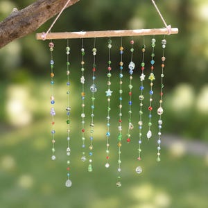 Op de afbeelding: Een hangende mobiel met een horizontale houten stok waaraan twaalf verticale draden zijn bevestigd. Elke draad is versierd met diverse veelkleurige glaskralen in groen, blauw, rood, geel en transparant, inclusief ster- en druppelvormige kralen.