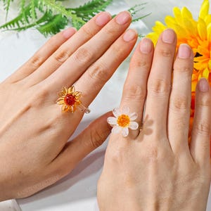 Puede incluir: Dos anillos ajustables con diseño de flor. El anillo de la izquierda tiene una flor amarilla y naranja con un centro rojo. El anillo de la derecha tiene una flor blanca con un centro amarillo y naranja.