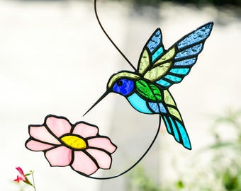 May include: A stained glass hummingbird with blue, green, and yellow wings hovers near a pink flower with a yellow center. The bird has a long, black beak and is suspended by a black metal wire, creating a decorative garden ornament.