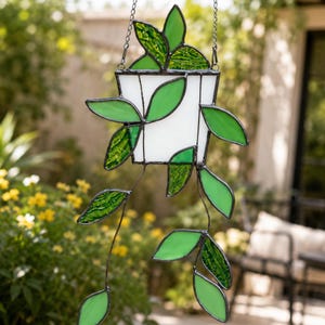 Puede incluir: Un adorno colgante de vidrieras que representa una planta con una maceta blanca trapezoidal y varias enredaderas con hojas de vidrio en distintos tonos de verde. La pieza está unida con líneas de soldadura metálica oscura y una cadena.