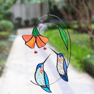 Puede incluir: Un atrapasoles de colibrí de vidrieras con una flor naranja y hojas verdes. Dos colibríes con detalles azules, rojos y blancos están posados en un alambre curvado. El atrapasoles cuelga de una cadena.