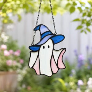 May include: A stained glass suncatcher shaped like a ghost wearing a blue witch hat. The ghost is white with light pink accents on the sides, and the hat features a dark blue band. It hangs from a thin black metal chain against a blurred background.