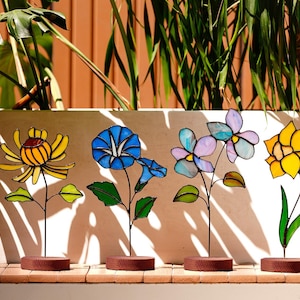 May include: A row of six colorful stained glass flower sculptures. Each flower has a unique design and color palette, including red, yellow, blue, and purple. The flowers are mounted on thin metal stems with brown circular bases.