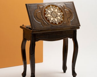 May include: A dark brown wooden table with a carved floral design on the top. The table has four legs and a small shelf underneath. The table is closed and appears to be made of wood.