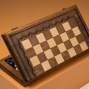 May include: A wooden chessboard with a hinged lid, revealing a light brown and dark brown checkerboard pattern. The board is decorated with a carved design and the letters A through H and numbers 1 through 8 are engraved on the board.
