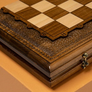 May include: A wooden chessboard with a hinged lid and a carved design. The board is closed and the lid is open. The board is made of dark wood and has a light wood inlay. The board is numbered 1 through 8 and lettered A through H.