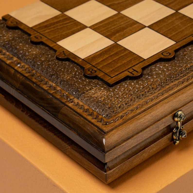May include: A wooden chessboard with a hinged lid and a carved design. The board is closed and the lid is open. The board is made of dark wood and has a light wood inlay. The board is numbered 1 through 8 and lettered A through H.