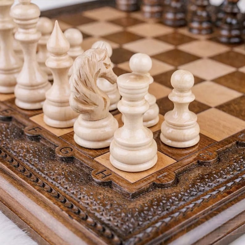 May include: A wooden chessboard with carved details and a set of white chess pieces, including a knight, pawn, and rook. The pieces are made of light wood and have a smooth finish.
