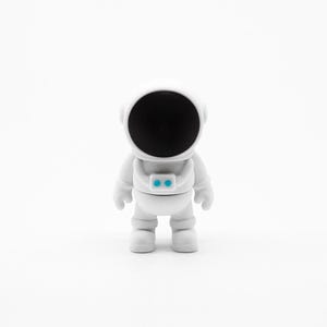 May include: A white astronaut figurine with a black circular face and two small blue lights on the chest. The astronaut is standing upright against a white background.