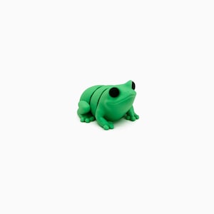 May include: A vivid green frog ornament with prominent black eyes. The frog features a smooth, curved body with segmented detailing and small feet. The ornament is set against a plain white backdrop, emphasizing its bright colour and simple design.