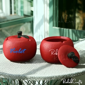 以下が含まれることがあります： 黒い茎と葉が付いた、リンゴ型の赤いセラミック容器。 1つは蓋付き、もう1つは開いています。 「Violet」の文字が青で、もう一方には「Rob」の文字が白で書かれています。 金属製のテーブルの上にあります。