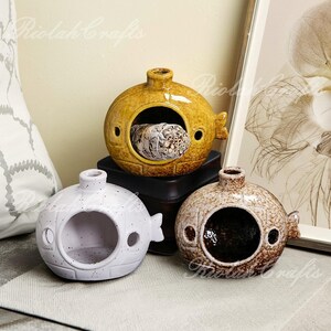 May include: Three ceramic submarine-shaped incense holders in yellow, white, and brown. The yellow holder contains a bundle of sage. Each holder has a circular opening and fin-like details. The holders are displayed on a table.