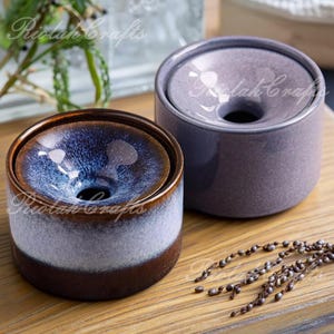 May include: Two ceramic containers with a unique design. One is a deep blue with brown and white accents, the other is a muted purple. Both have a central hole and a lid. The containers are on a wooden surface.