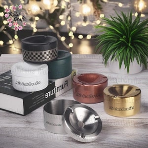 Könnte beinhalten: Ein Set aus sechs verschiedenen Aschenbechern in verschiedenen Farben und Materialien, darunter Schwarz, Weiß, Grün, Kupfer, Gold und Edelstahl. Die Aschenbecher sind alle rund und haben eine tiefe Vertiefung zum Auffangen von Zigarettenstummeln. Die Aschenbecher sind auf einem Holztisch mit einer Pflanze im Hintergrund angeordnet.
