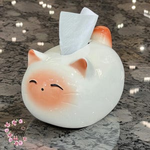 May include: White ceramic tissue box holder shaped like a cat with orange accents. The cat has closed eyes and a smiling expression. A white tissue is pulled out of the box.