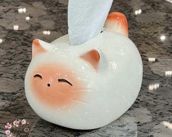 Puede incluir: Porta pañuelos de cerámica blanca con forma de gato con detalles en naranja. El gato tiene los ojos cerrados y una expresión sonriente. Un pañuelo blanco se saca de la caja.
