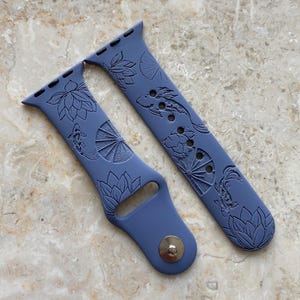 May include: Two blue watch bands with a detailed design of koi fish and lotus flowers. The bands are made of a flexible material and have a silver metal clasp. The bands are designed to fit a smartwatch.