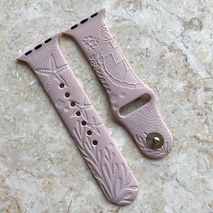 Può includere: Due cinturini per orologio rosa chiaro con disegni incisi a tema oceanico. Uno presenta stelle marine e alghe, l'altro una tartaruga marina e coralli. I cinturini sono realizzati in materiale flessibile e hanno una chiusura in metallo.