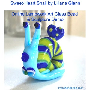 Puede incluir: Una cuenta de vidrio de caracol azul y verde con una concha en forma de corazón. El caracol tiene un cuerpo rayado azul y blanco y una cabeza verde. La cuenta es una escultura de vidrio de arte lampwork.