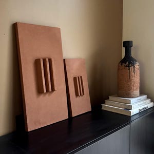 Può includere: Due opere d'arte da parete rettangolari color terracotta con linee verticali in rilievo e una bottiglia strutturata con vernice nera che cola. Gli oggetti sono esposti su un mobile nero, con libri impilati sopra.