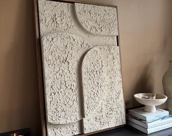Puede incluir: Arte mural abstracto beige texturizado en un marco de madera oscura. La obra de arte presenta un diseño en capas con formas curvas y rectangulares. La pieza se exhibe sobre un mueble negro, con libros y una vela cerca.