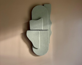 May include: A sage green abstract wall sculpture composed of layered geometric shapes. The artwork features a combination of rectangular, curved, and irregular forms, creating a three-dimensional effect. The sculpture is mounted on a neutral-toned wall.