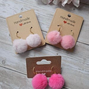 Könnte beinhalten: Drei Paar handgefertigte Pompon-Ohrringe in Rosa- und Weißtönen, präsentiert auf beigefarbenen Karten. Auf den Karten steht "Handmade with love". Die Ohrringe haben goldfarbene Haken.