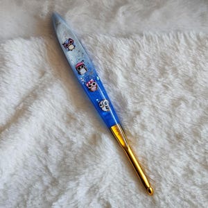 Peut inclure: Un crochet bleu et doré avec un manche décoratif. Le manche est orné de motifs de hiboux et de paillettes, et le crochet est de couleur or. Le crochet mesure environ 15 cm de long.