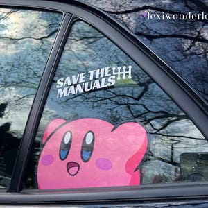 May include: A car window sticker featuring a pink Kirby character with blue eyes and a smiling expression. Above Kirby is a white sticker with the text "SAVE THE MANUALS". The background shows a view of trees and sky.