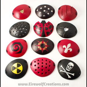 An assortment of handmade leather eye patches with various designs, including rivets, scarred, ladybug, spiral, fleur de lis, radiation hazard, Jolly Roger, see-through hearts, see-through perforated, and see-through compass rose.