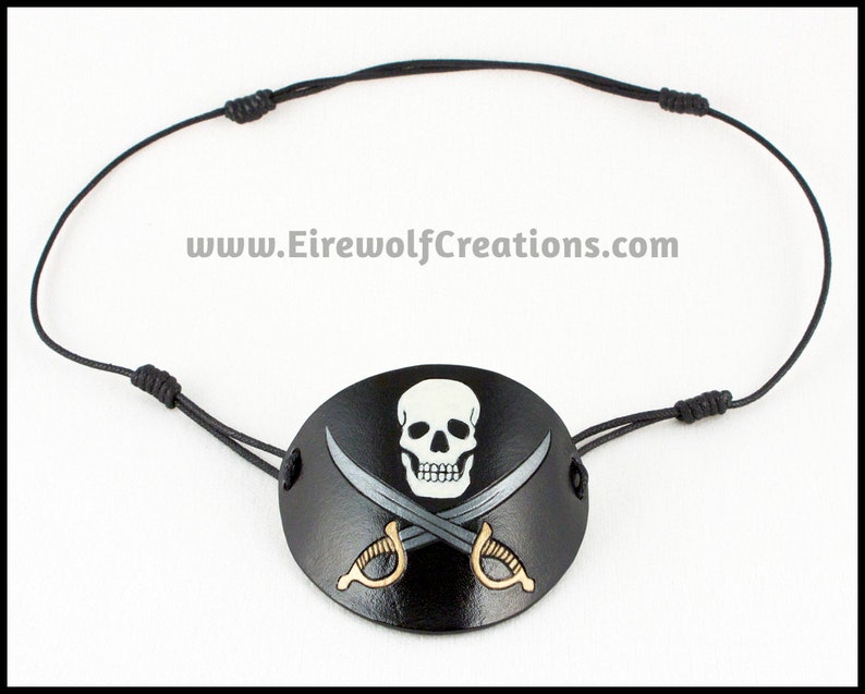 Handmade leather eyepatch with a Jolly Roger skull-and-crossed-swords design carved and painted on the leather. The skull is bone-white on black, and the swords are painted silver with golden hilts. Black cotton cords with sliding knots.