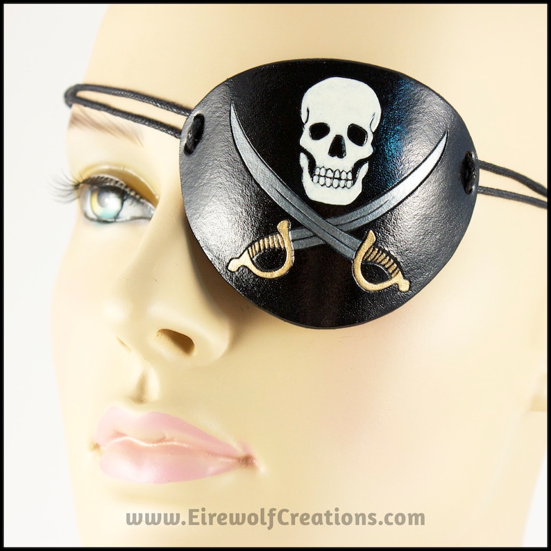 Handmade leather eyepatch with a Jolly Roger skull-and-crossed-swords design carved and painted on the leather. The skull is bone-white on black, and the swords are painted silver with golden hilts.