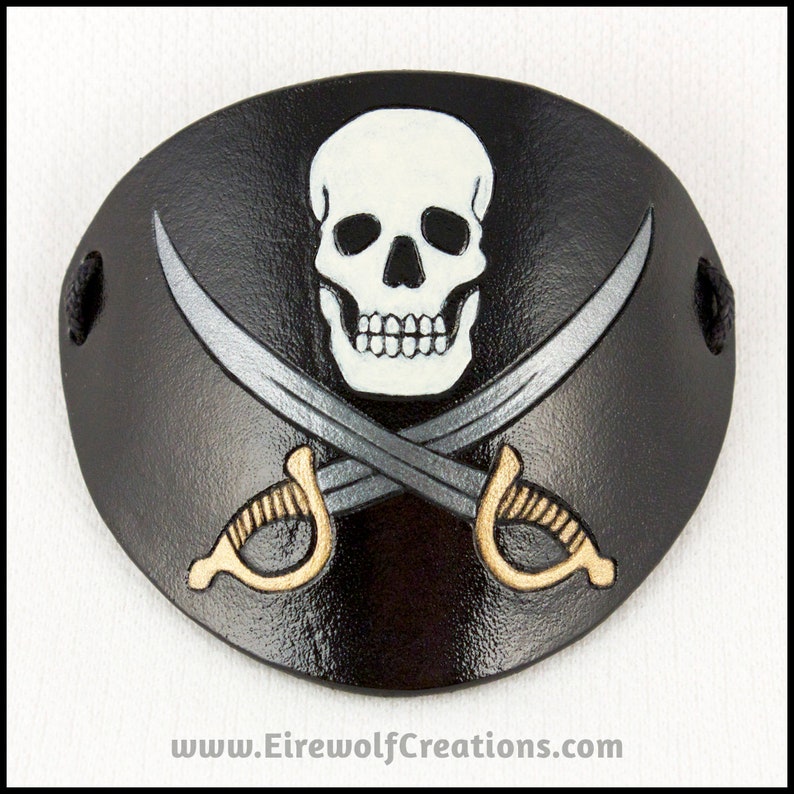 Handmade leather eyepatch with a Jolly Roger skull-and-crossed-swords design carved and painted on the leather. The skull is bone-white on black, and the swords are painted silver with golden hilts.