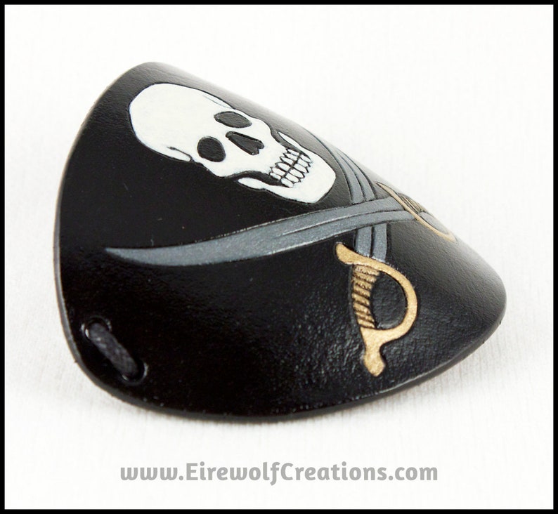 Handmade leather eyepatch with a Jolly Roger skull-and-crossed-swords design carved and painted on the leather. The skull is bone-white on black, and the swords are painted silver with golden hilts. Side view.