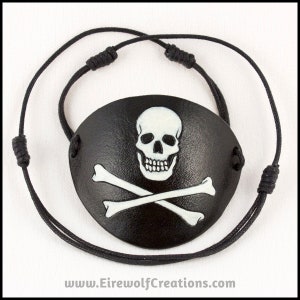 Handmade leather eyepatch with a skull-and-bones Jolly Roger carved into the leather and painted bone-white on a black background. Black cotton cords with adjustable sliding knots.
