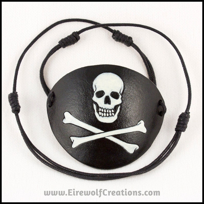 Handmade leather eyepatch with a skull-and-bones Jolly Roger carved into the leather and painted bone-white on a black background. Black cotton cords with adjustable sliding knots.