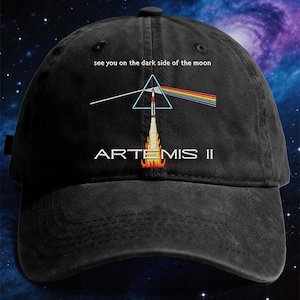 May include: Black baseball cap with the text "see you on the dark side of the moon." The cap features a graphic of a rocket launching through a prism, creating a rainbow. The text "ARTEMIS II" is embroidered below the rocket.