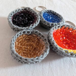Peut inclure: Quatre porte-clés crochetés avec des anneaux argentés. Chaque porte-clés présente un motif circulaire rempli de perles colorées. Les couleurs des perles comprennent le noir, le bleu, l'or et le rouge. La bordure au crochet est grise.